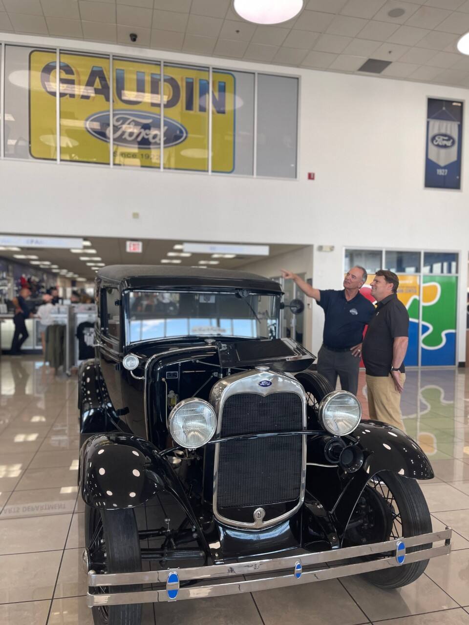Two men discuss a vintage black Ford vehicle in a dealership showroom.
